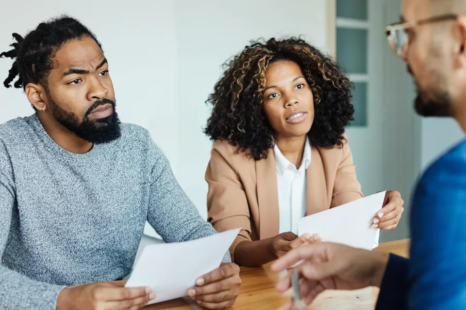 A Black couple holding contracts sits across the desk from a white mortgage lender.